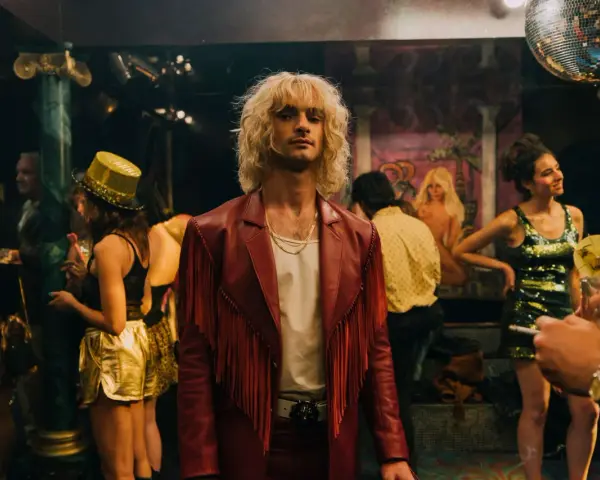 Ein Mann mit blonden Haaren und rotem Fransen-Sakko steht im Mittelpunkt, umgeben von einer bunten **Besetzung von Luden** in einem Nachtclub mit Discokugel.