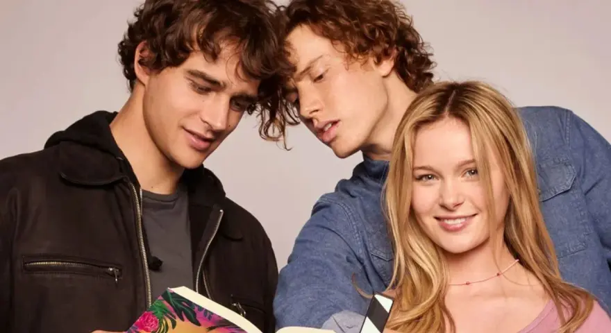 Die "Love Me, Love Me" Besetzung: Zwei junge Männer lesen ein Buch, während eine junge Frau lächelnd in die Kamera blickt.