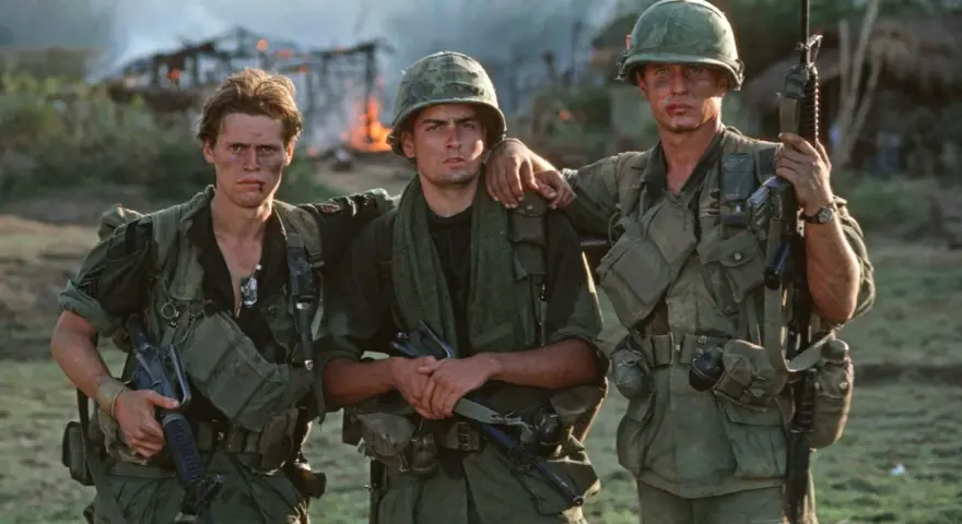 Die Besetzung von Platoon: Drei Soldaten in Uniform, mit Waffen und Ausrüstung, stehen vor brennenden Gebäuden.