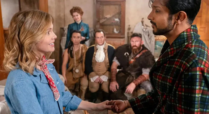 Ein Mann im karierten Bademantel hält die Hand einer Frau. Im Hintergrund sind mehrere historische Figuren zu sehen, die auf ein neues Abenteuer in "Ghosts Staffel 5" hindeuten.