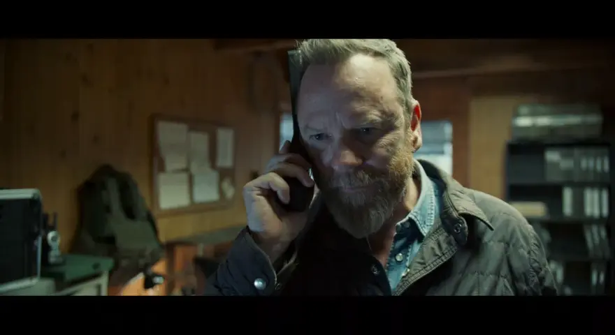 Mann mit Bart telefoniert, im Hintergrund eine Pinnwand und Regale. Besetzung von The Contractor.