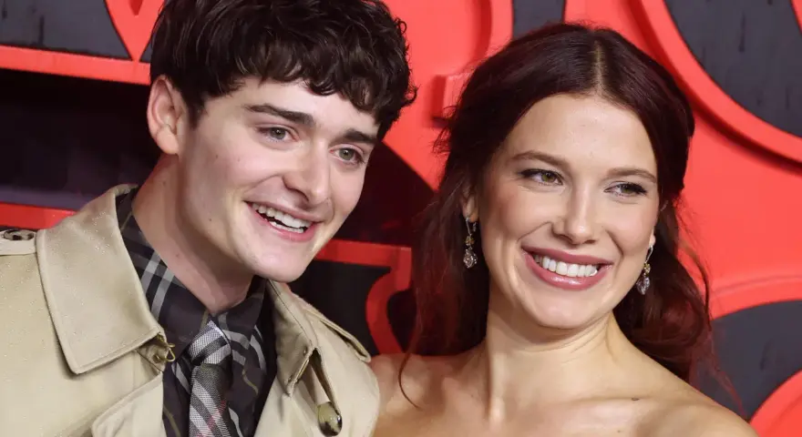 Noah Schnapp und eine Frau lächeln vor einem roten Hintergrund. Er trägt einen Trenchcoat und eine karierte Krawatte, wie man sie aus seinen Filmen kennt.