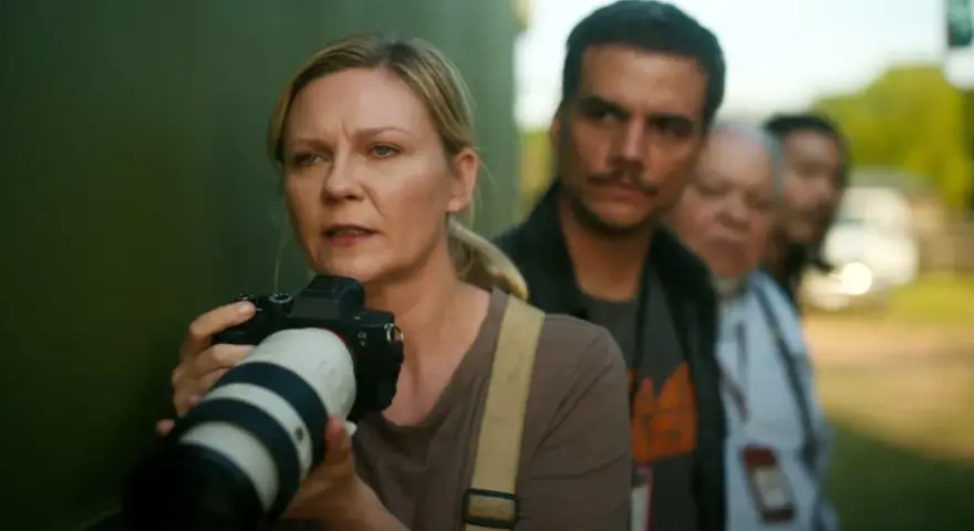 Kirsten Dunst mit Kamera, Teil der besetzung von Civil War, im Hintergrund unscharfe Personen.