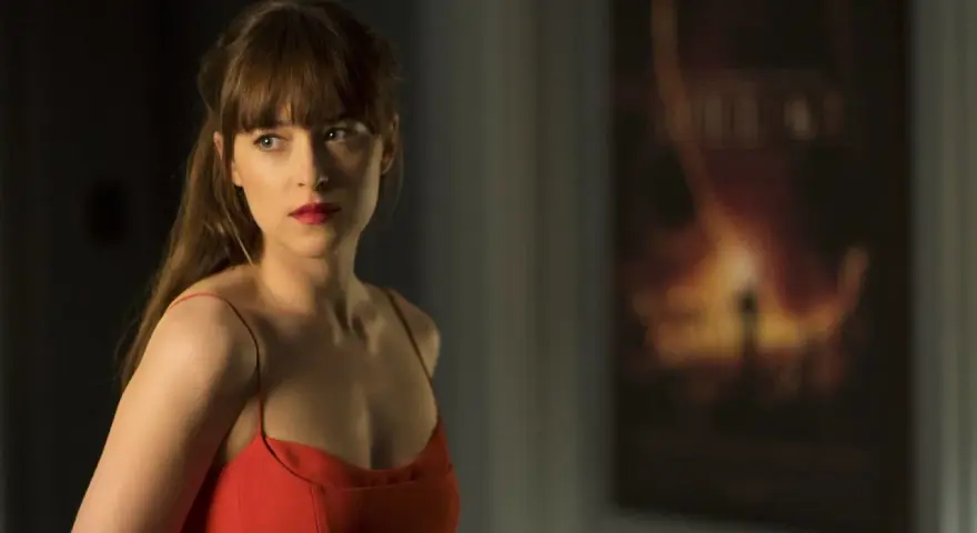 Dakota Johnson in einem roten Kleid, nachdenklich blickend. Ein Poster im Hintergrund deutet auf ihre Filme hin.