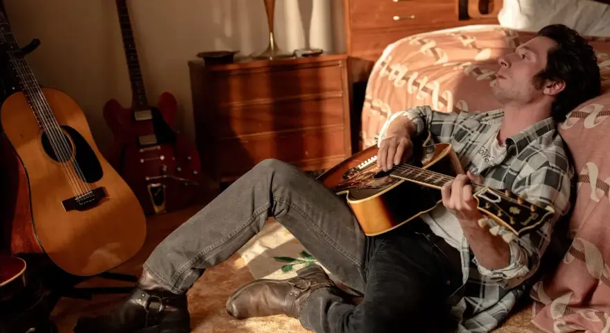 Ein Mann mit Gitarre, inspiriert von Bruce Springsteen, sitzt im Zimmer. Mehrere Gitarren stehen im Hintergrund.