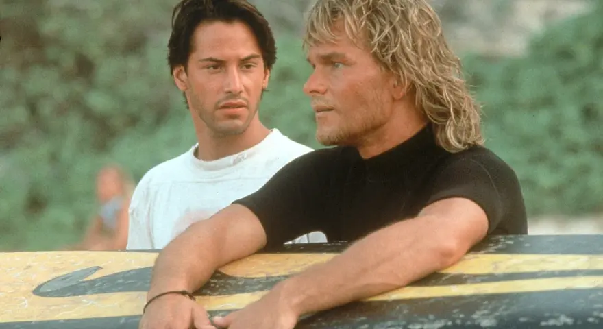 Keanu Reeves und Patrick Swayze, die Hauptdarsteller der Besetzung von Point Break, lehnen an einem Surfbrett.