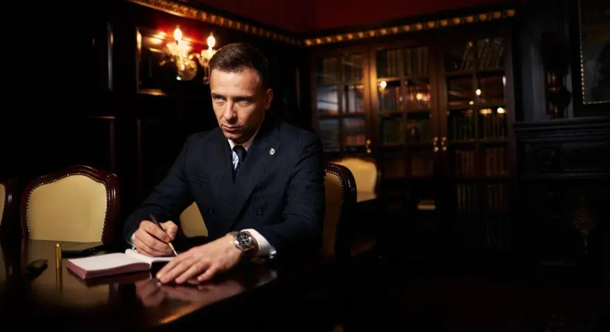 Ein Mann im Anzug schreibt in einem Notizbuch. Er könnte Teil der **Besetzung von The Lincoln Lawyer** sein, mit einem Bücherregal im Hintergrund.
