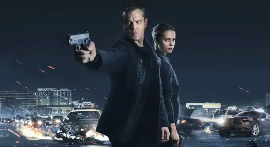 Jason Bourne und Alicia Vikander in Aktion. Ein Auto brennt, während Jason eine Waffe abfeuert. Die besetzung von Jason Bourne ist bereit für den Kampf.