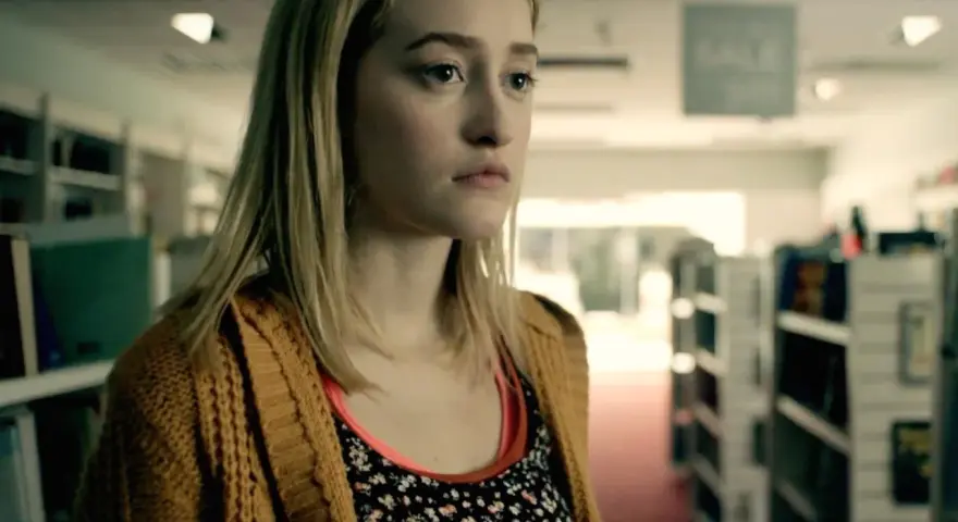 Junge Frau mit blonden Haaren und einem gelben Strickcardigan steht in einem Gang mit Bücherregalen. Ein Ausschnitt aus Gus Birneys Filme und Serien.