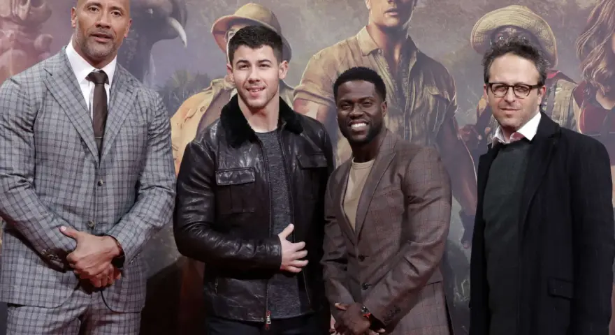 Die Besetzung von Jumanji: Dwayne Johnson, Nick Jonas, Kevin Hart und Jake Kasdan posieren vor einem Dschungel-Hintergrund.
