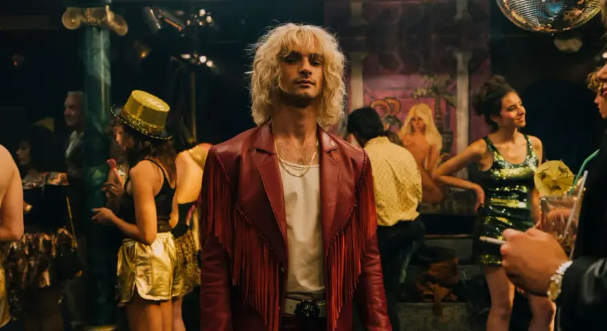 Ein Mann mit blonden Haaren und rotem Fransen-Sakko steht im Mittelpunkt, umgeben von einer bunten **Besetzung von Luden** in einem Nachtclub mit Discokugel.