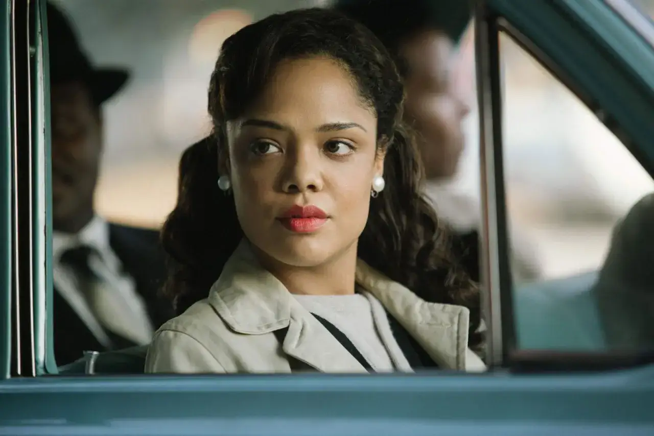 Tessa Thompson in einem Auto, ein Ausschnitt aus ihren Filmen. Sie trägt einen beigen Mantel und Perlenohrringe.