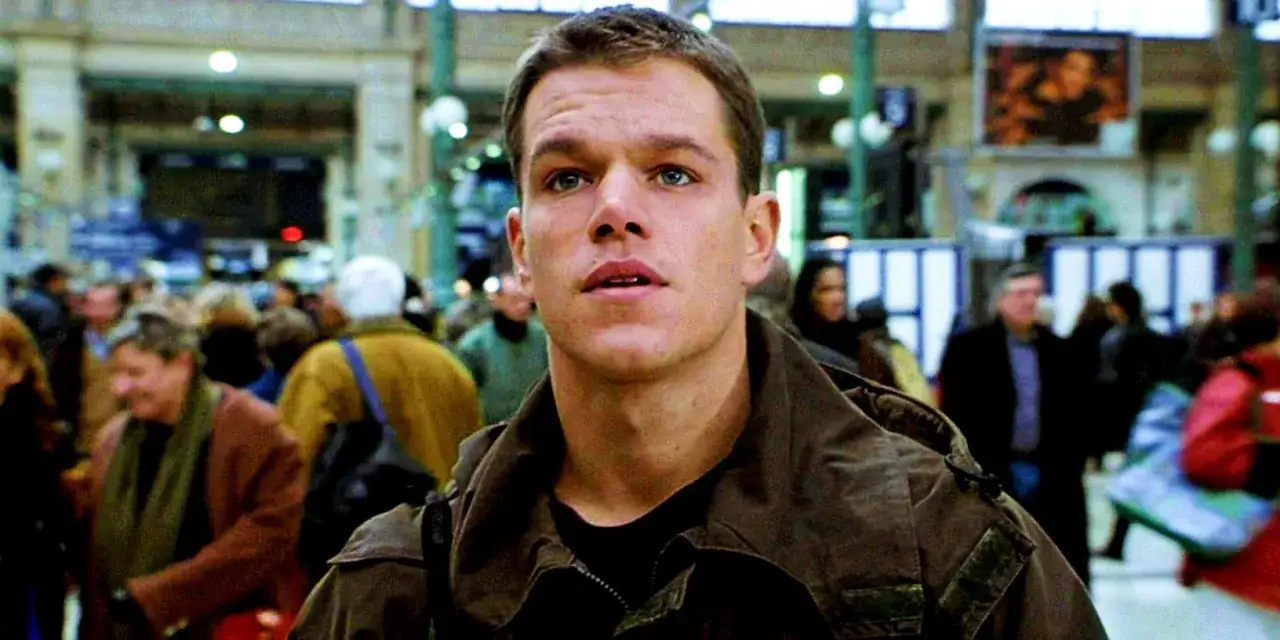 Matt Damon als Jason Bourne in einer belebten Bahnhofshalle. Die Besetzung von Jason Bourne zeigt ihn nachdenklich inmitten der Menge.