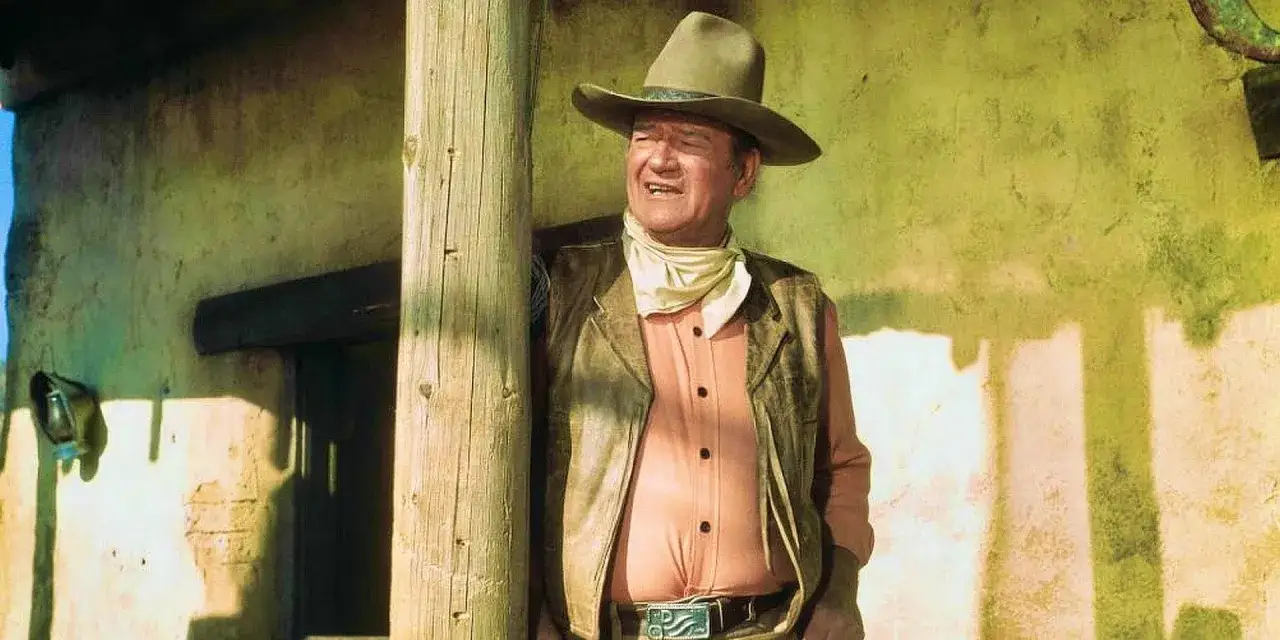 John Wayne in seiner Rolle bei der besetzung von Rio Lobo, lehnt an einem Holzpfosten vor einer verwitterten Wand.