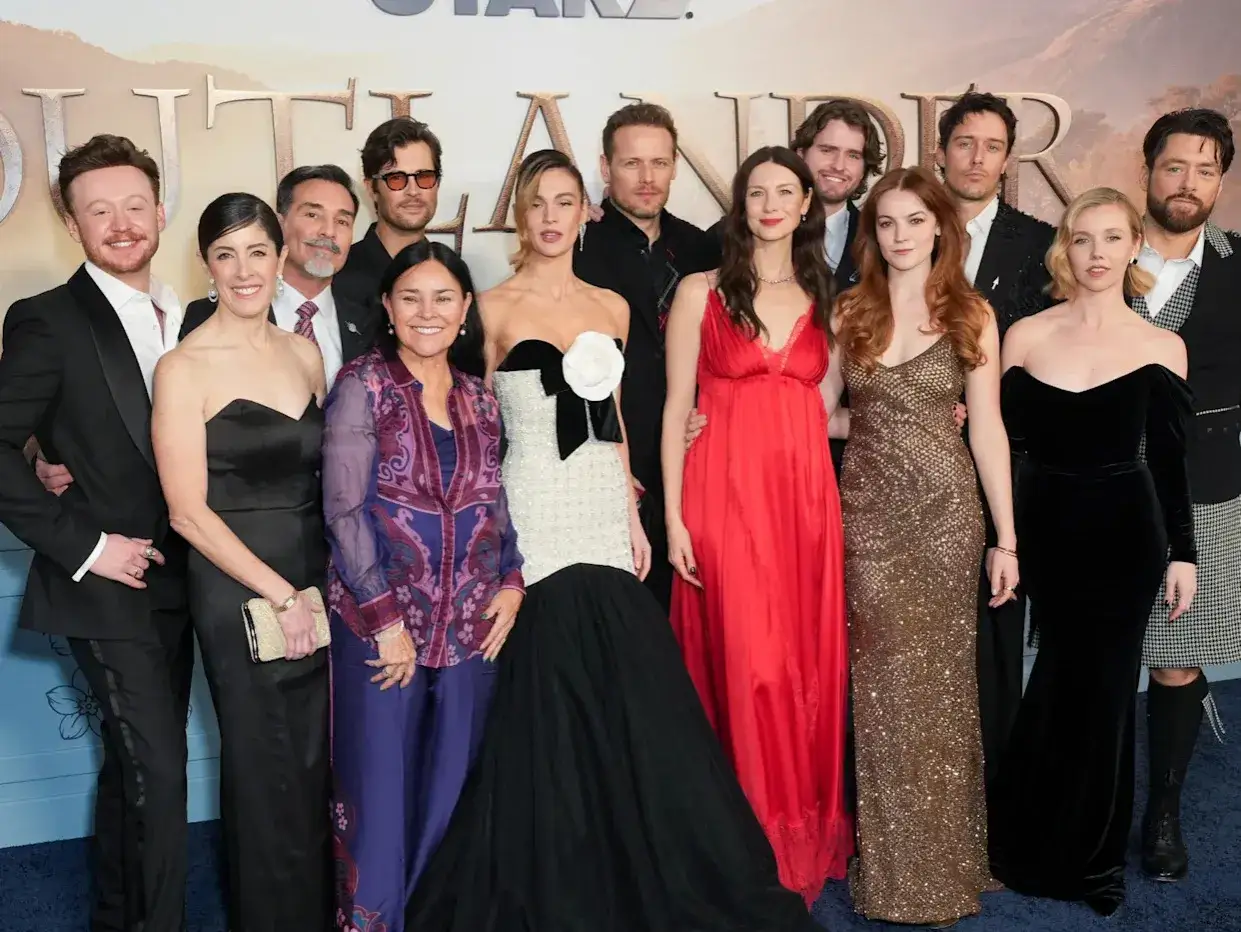 Die Besetzung von Outlander posiert für ein Gruppenfoto. Die Schauspieler tragen elegante Kleidung und lächeln in die Kamera.