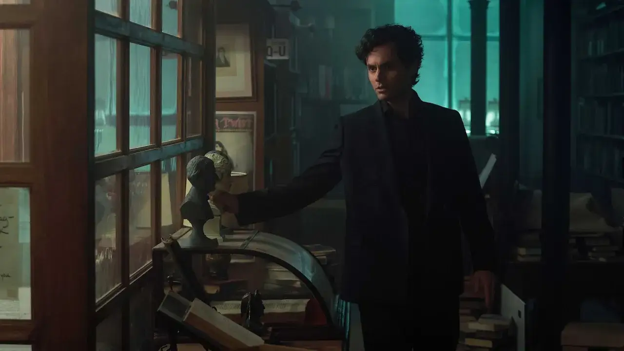 Joe in dunklem Anzug in einer Bibliothek, die auf eine Büste zeigt. Staffel 5 von YOU.