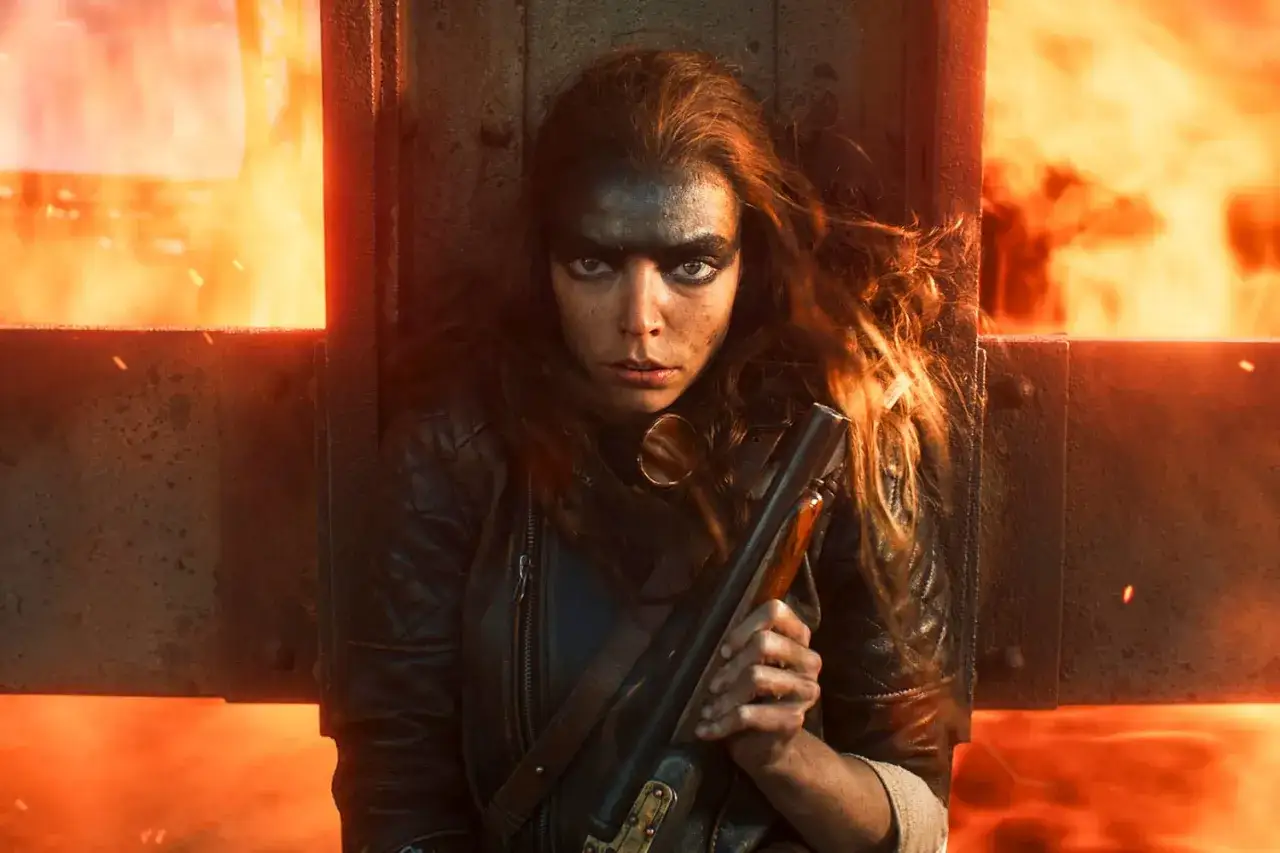 Furiosa: A Mad Max Saga. Eine junge Frau mit Kriegsbemalung und einem Gewehr blickt entschlossen in die Kamera, umgeben von Flammen.