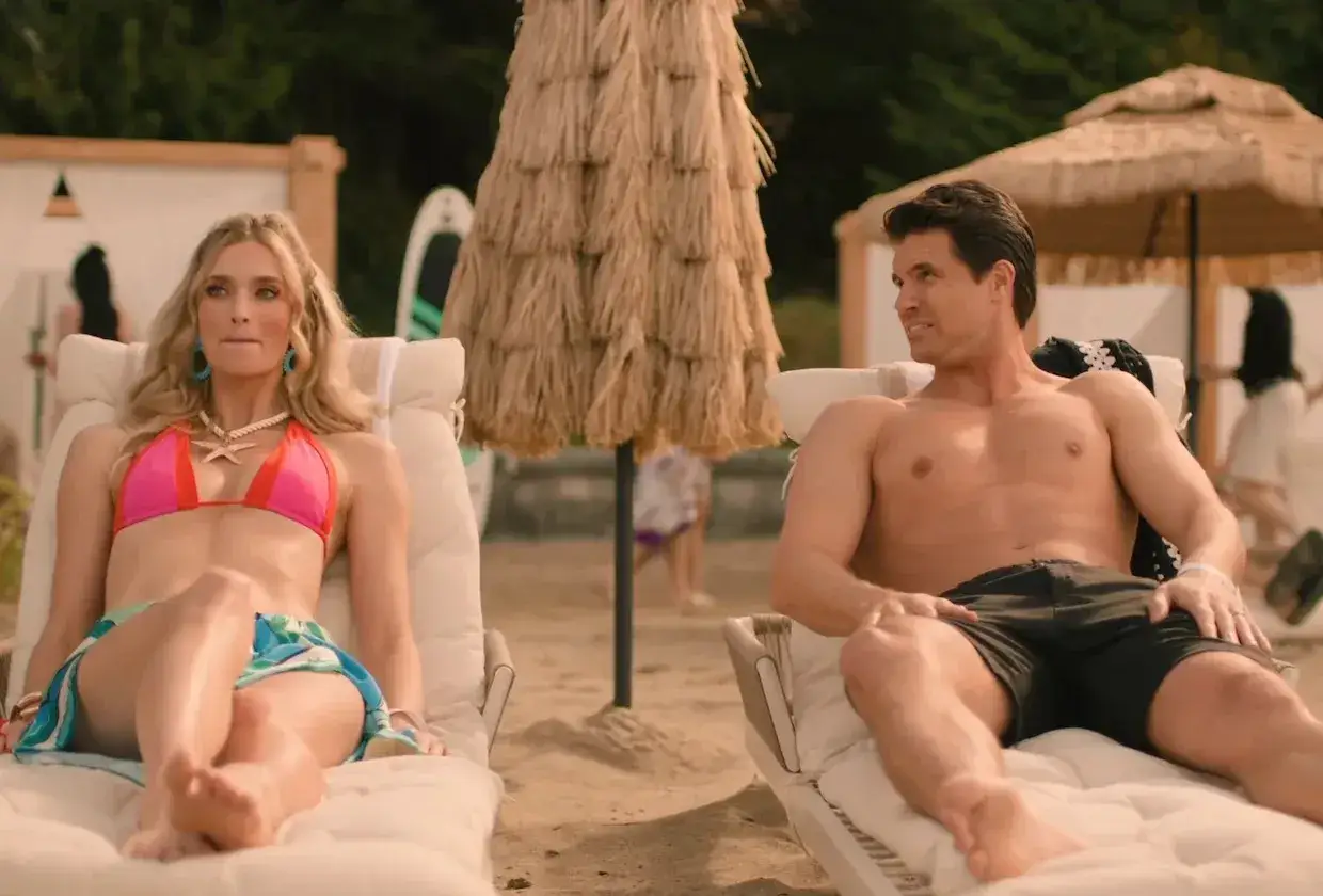 Ein Paar entspannt am Strand. Sie trägt einen pinken Bikini, er kurze Hosen. Vielleicht ist das die upload staffel 4?