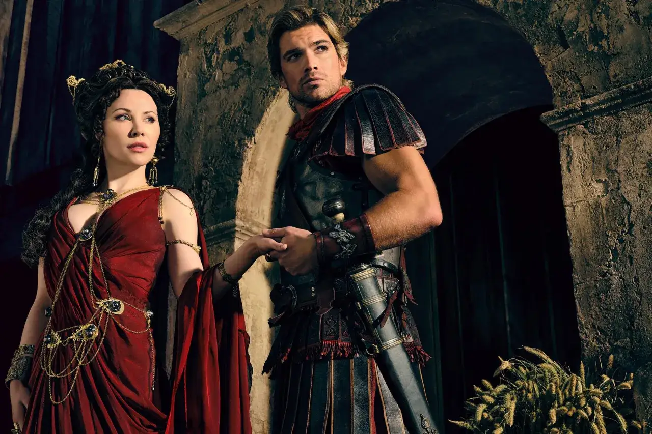 Die Besetzung von Spartacus: Ashur und Lucretia, in prunkvollen Gewändern und Rüstung, halten Händchen vor einer antiken Kulisse.