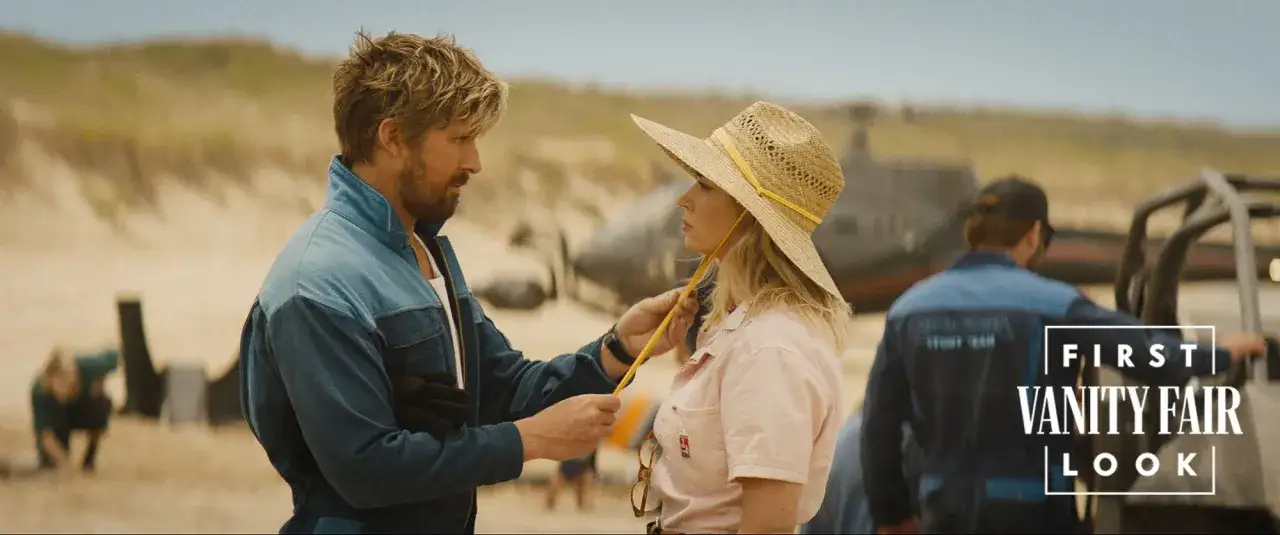 Ein Mann im Blaumann spricht mit einer Frau mit Strohhut am Strand. Er ist der