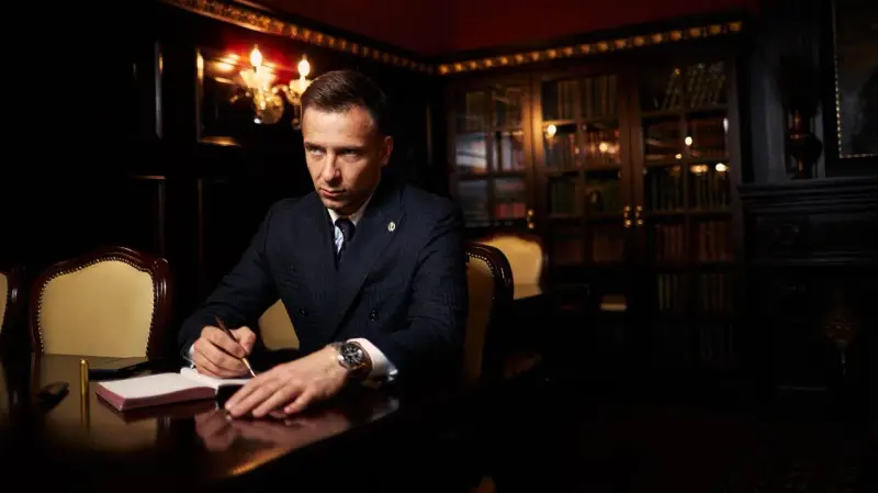 Ein Mann im Anzug schreibt in einem Notizbuch. Er könnte Teil der **Besetzung von The Lincoln Lawyer** sein, mit einem Bücherregal im Hintergrund.